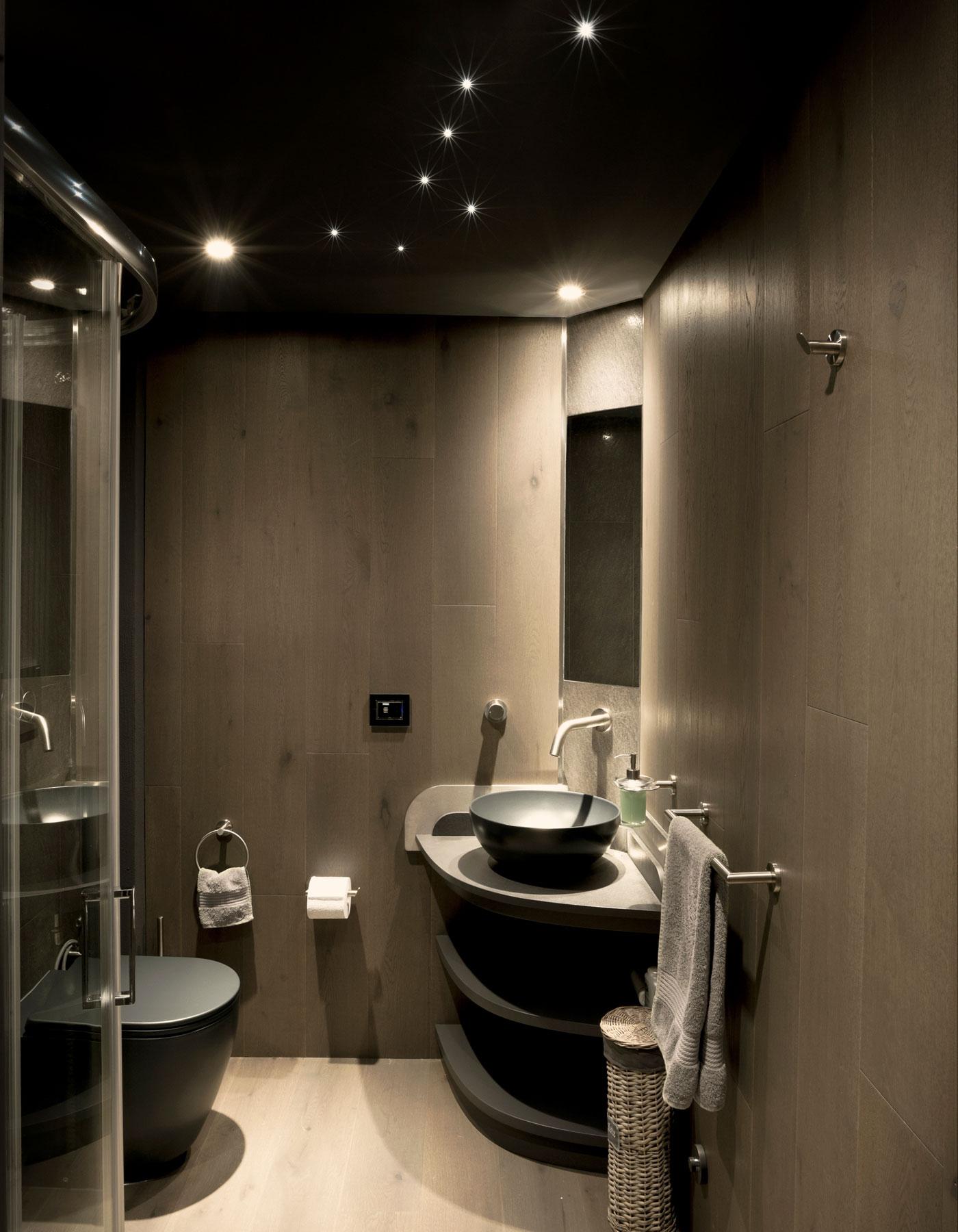 Bagno moderno rivestito in legno grigio con lavabo angolare, illuminazione con faretti spot e soffitto scuro