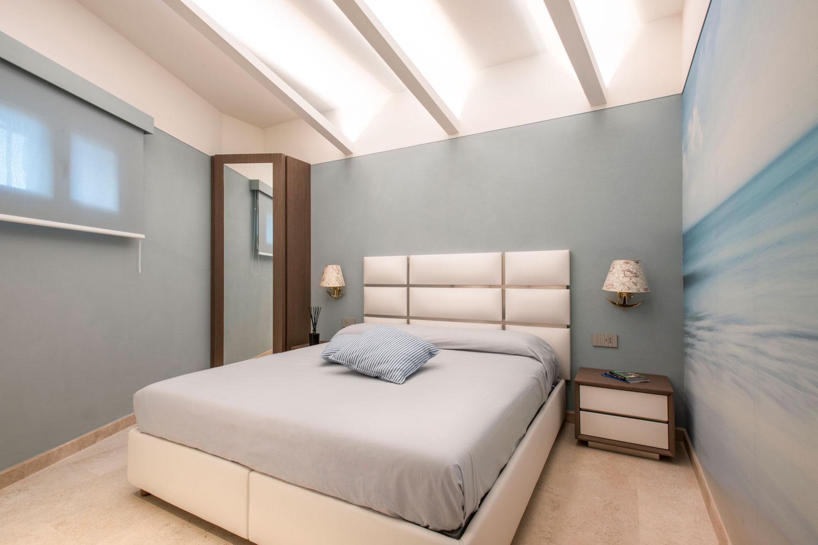 Camera da letto con pareti azzurre, testata in ecopelle bianca e finiture acciaio, armadio ad angolo con specchio