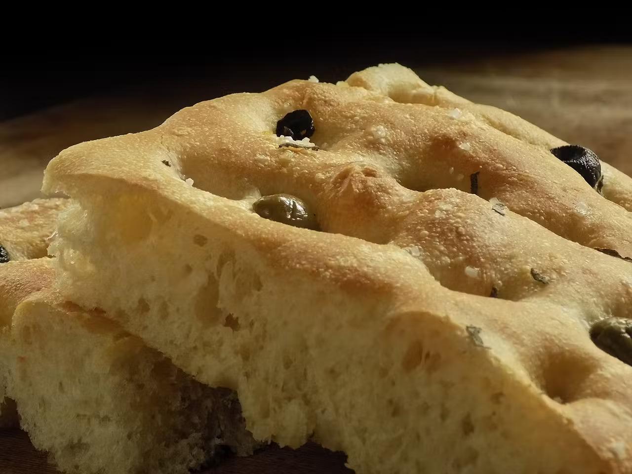 Focaccia di farro bianco alta e soffice, con una superficie dorata e croccante, cosparsa di rosmarino fresco e olive, tagliata in quadrati pronti per essere serviti