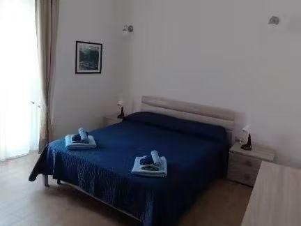 Camera da letto dell'appartamento al primo piano, letto matrimoniale dotato di asciugamani, luci e prese vicino al letto, stanza luminosa ma dicreta, porta finestra oscurabile grazie alle tende e si affaccia su una strada tranquilla di un piccolo paese del centro Sardegna.