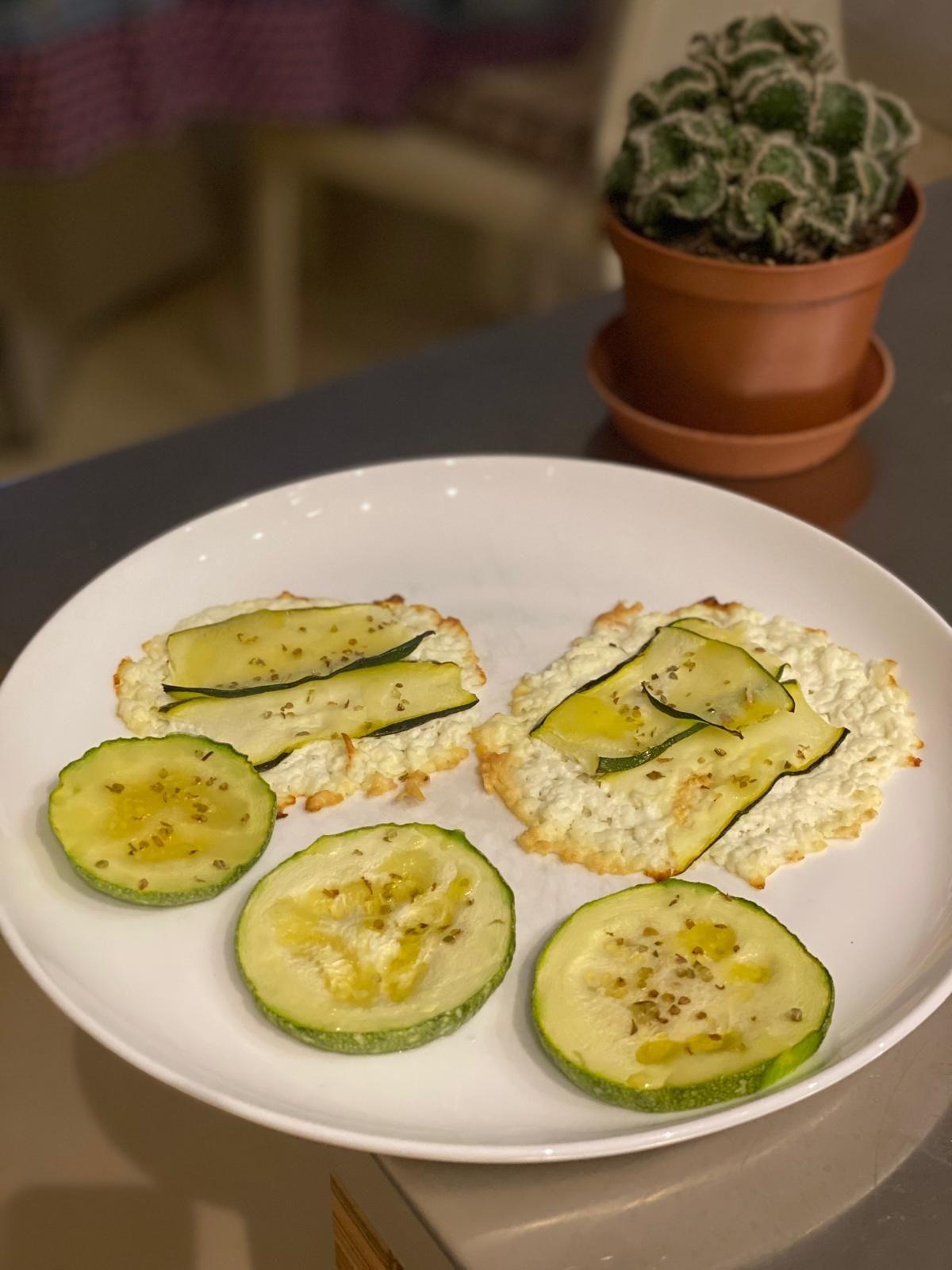 Ingredienti: 200 grammi di feta greco di capra, zucchine, origano, olio extravergine d’oliva q.b. Procedimento: Grattugiare la feta con una grattugia a fori grossi e, con un cucchiaio, distribuirla in tanti mucchietti su una teglia rivestita con carta forno. Sistemare ogni mucchietto in modo che abbia una forma circolare e piatta. Lavare e tagliare le zucchine a fettine sottili e porle sopra la feta. Condire con origano e un filo d’olio evo. Cuocere in forno a 180°C per circa 15-20 minuti, fino a far sciogliere la feta e cuocere le zucchine. Negli ultimi 5 minuti, usare il grill. Varianti: la feta di capra può essere sostituita da quella di latte vaccino e possono essere utilizzate altre verdure al posto delle zucchine, condite con altre erbe aromatiche.